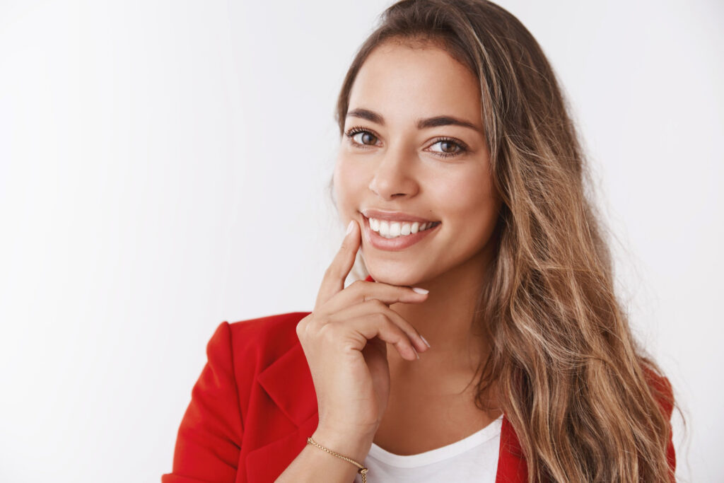 Mulher sorridente com dentes brancos alinhados, vestindo blazer vermelho, tocando o queixo em reflexão sobre lentes de contato dental em Perdizes.