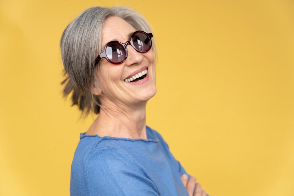 Mulher grisalha de óculos escuros sorrindo em fundo amarelo. Exemplo de prótese protocolo em Perdizes na Saraiva Odontologia